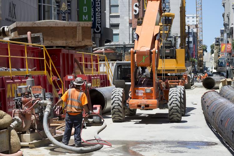 Construction worker operating machinery on Stockton Street with large pipes and equipment visible.