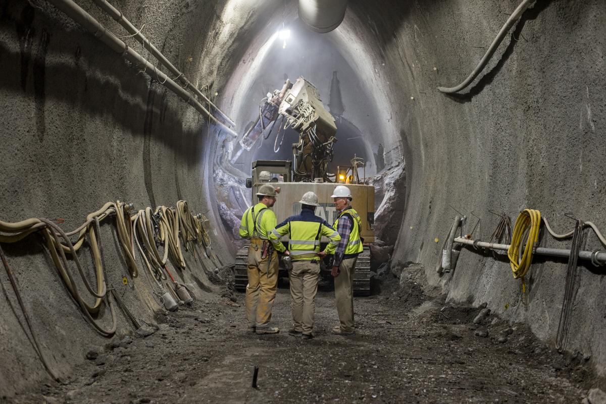 Workers in hard hats and safety vests stand in a large tunnel with an excavator and construction equipment visible in the distance.