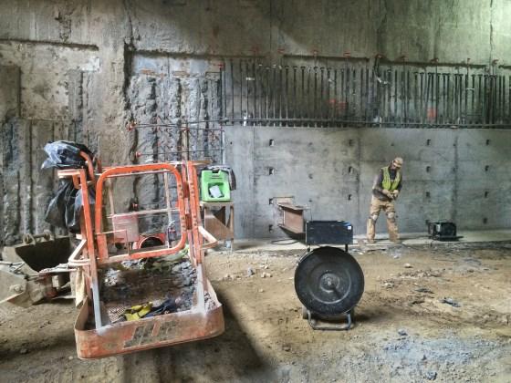 Construction workers and equipment inside underground station excavation site with concrete walls and support structures.