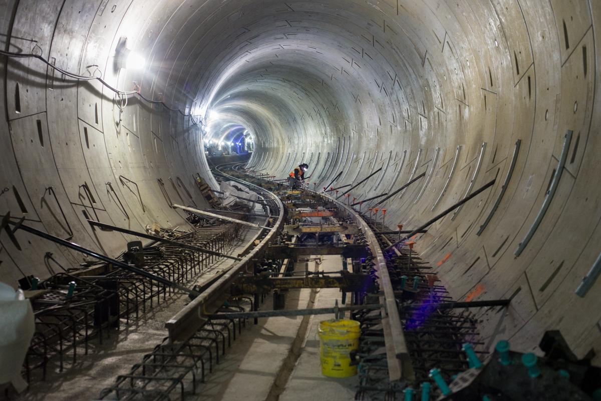 Underground tunnel under construction with railway tracks, lighting, and workers in safety gear.
