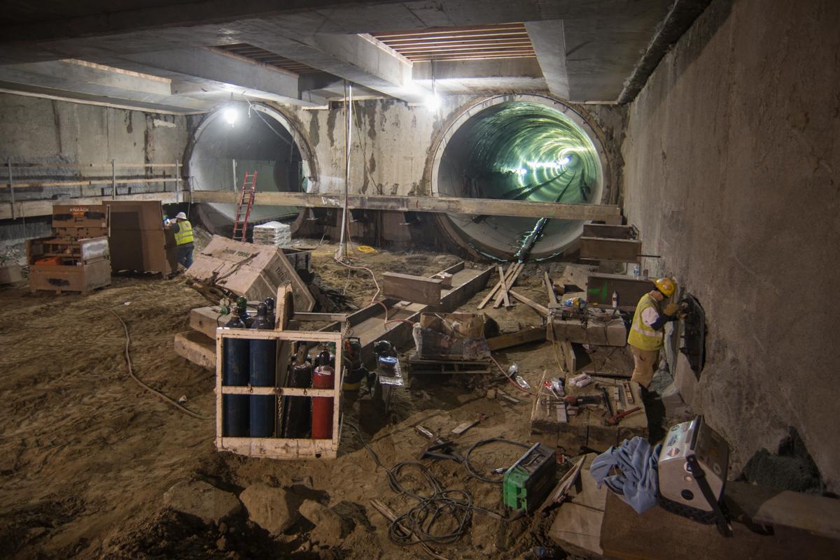 Construction workers install temporary steel bracing inside underground transit station tunnel with large circular pipes.