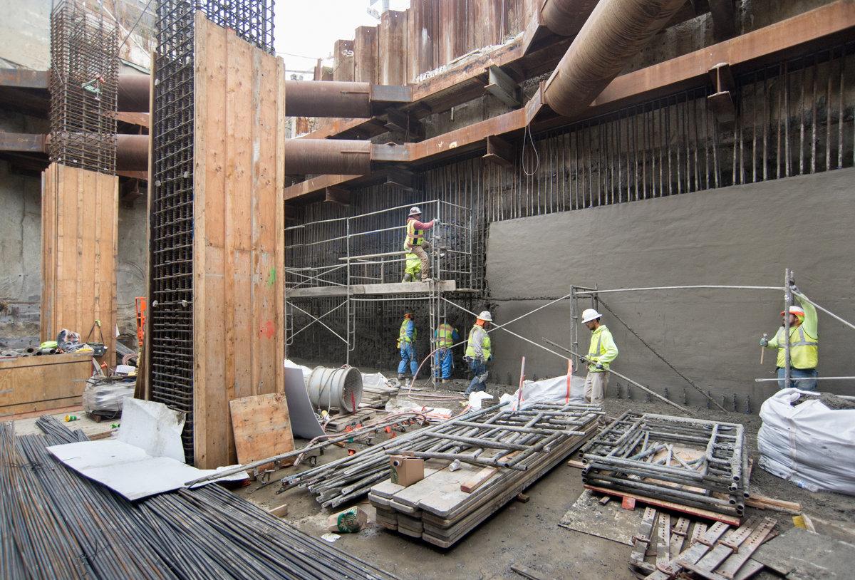 Construction workers disassemble scaffolding at underground transit station with reinforced concrete wall and steel structural elements visible.