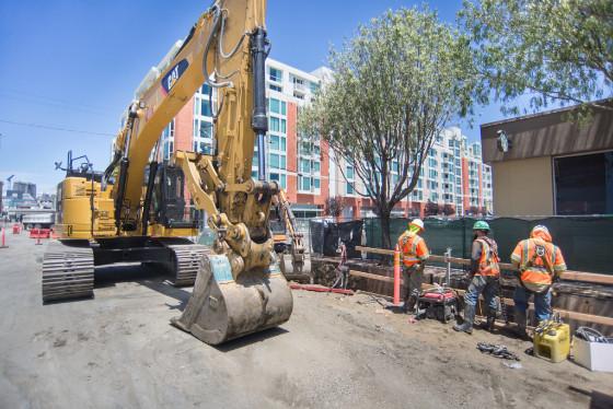 Yellow excavator with bucket operates at construction site with workers in orange safety vests nearby.