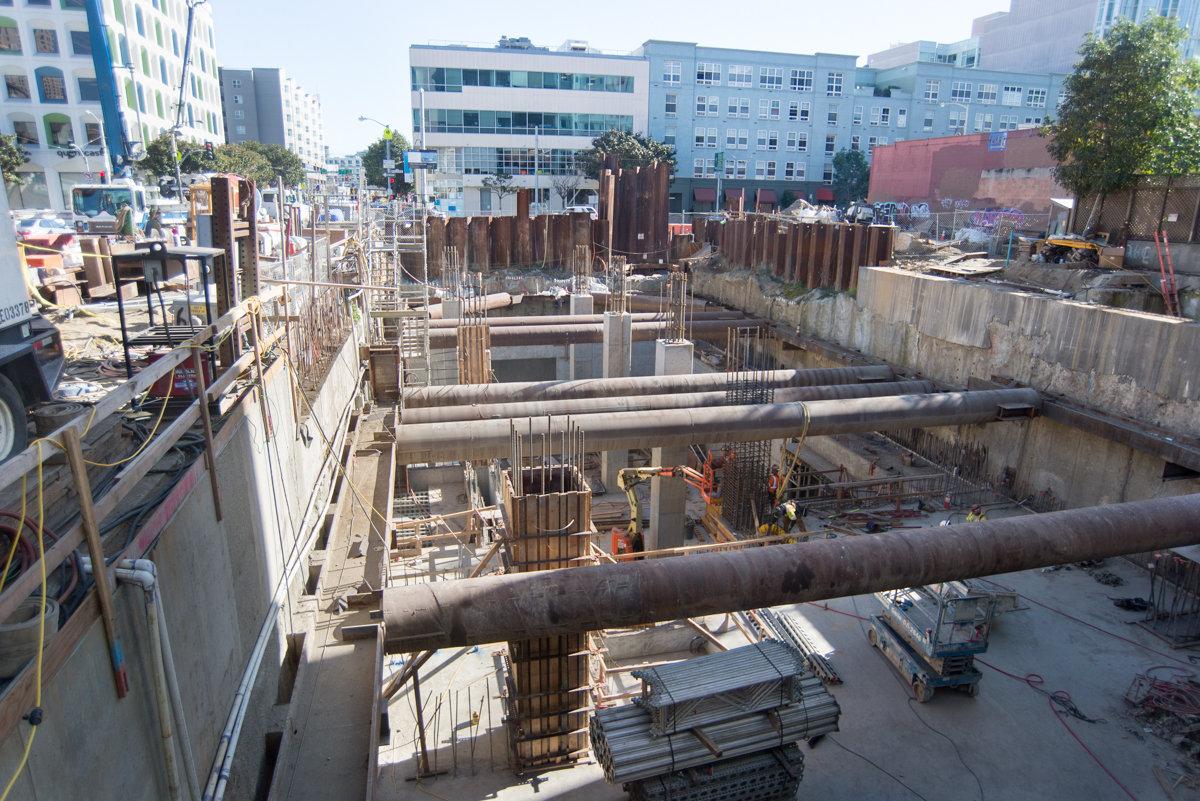Central Subway construction site showing excavated station pit with steel reinforcement, shoring walls, and surrounding San Francisco buildings.
