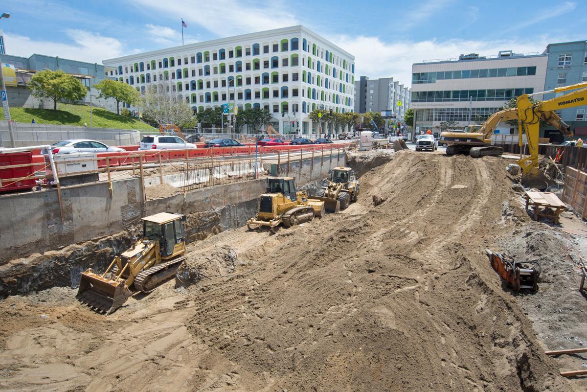 Active excavation site on 4th Street with heavy equipment, dirt mounds, and red safety barriers surrounding construction area near downtown buildings.