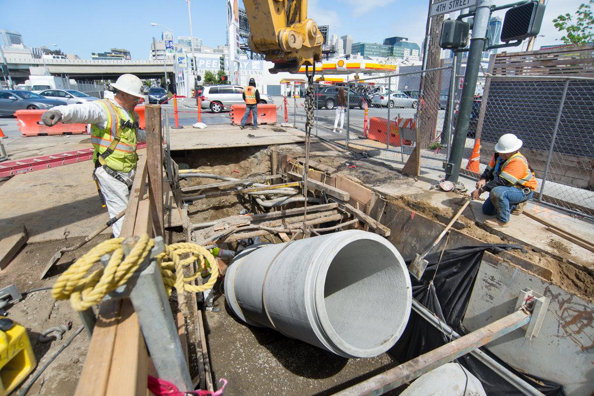 Construction workers lower large concrete pipe into excavation pit for Central Subway project on 4th Street.