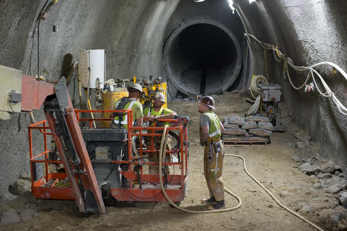 Workers in safety gear operate equipment inside a large concrete tunnel during sewer excavation work.