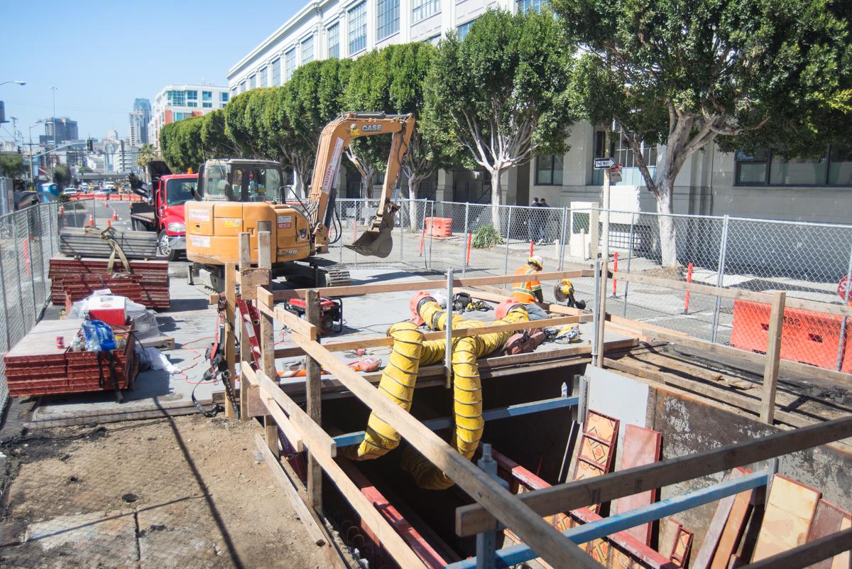 Construction crew working in excavated pit with yellow safety equipment and orange excavator on urban street.