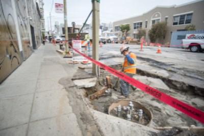 Construction worker in safety vest at underground utility excavation site on city street with red barriers.