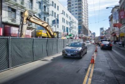Construction site on Stockton Street with yellow excavator, protective fencing, and single lane of traffic in each direction.