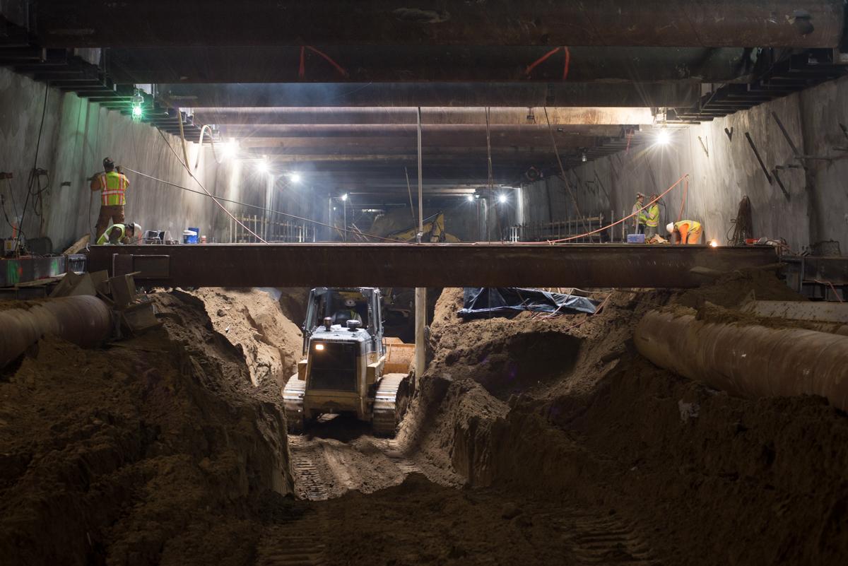 Underground transit station excavation with temporary steel bracing installed under Stockton Street, workers and equipment visible.