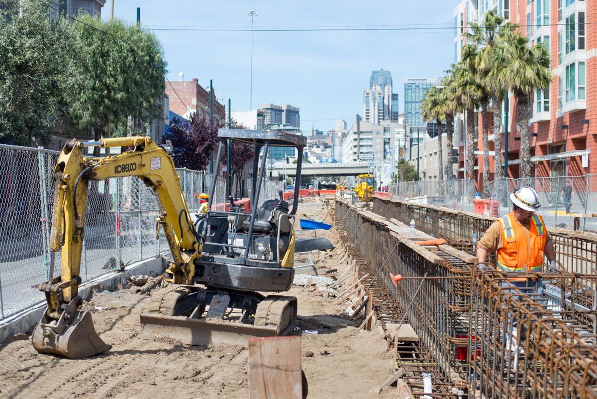 Construction crew operating excavator on transit track work site with city skyline in background.