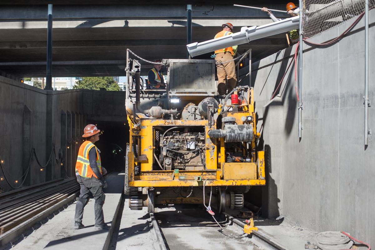 Workers operate a concrete pump truck to deliver concrete into a hopper car inside a tunnel portal.