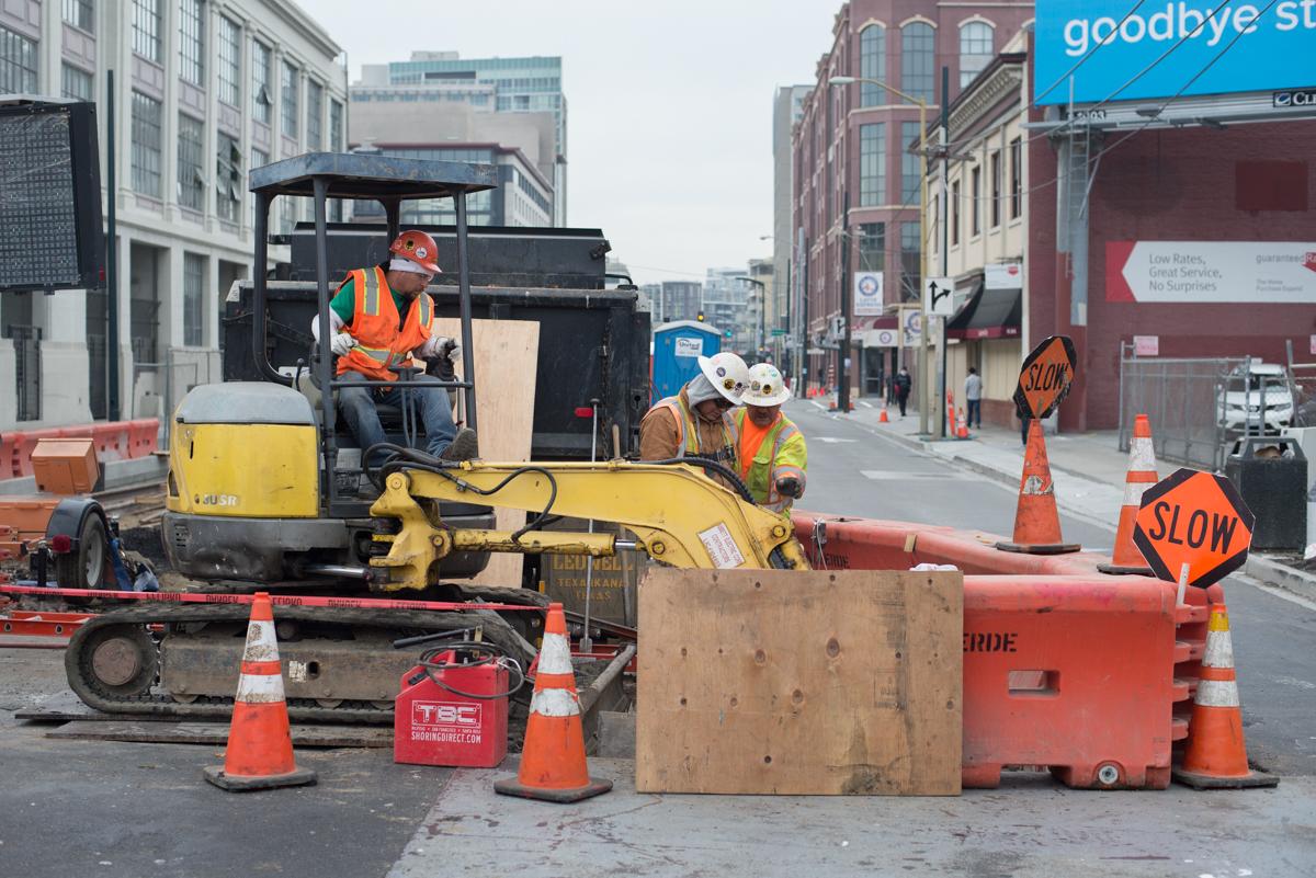 Construction crew operating equipment and installing utilities on city street with traffic control barriers.