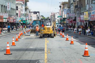 Construction equipment and traffic cones block Stockton Street during utility work, with buildings and pedestrians visible along the urban street.