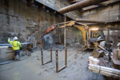 Construction workers operate jackhammer and excavator equipment inside an underground station cavern during preparation work.