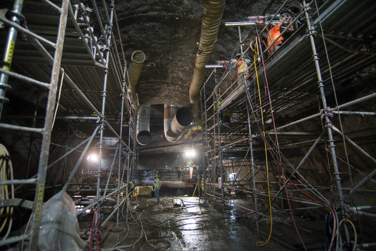 Workers on scaffolding drill pilot holes inside underground tunnel construction site with barrel vault ceiling and equipment.