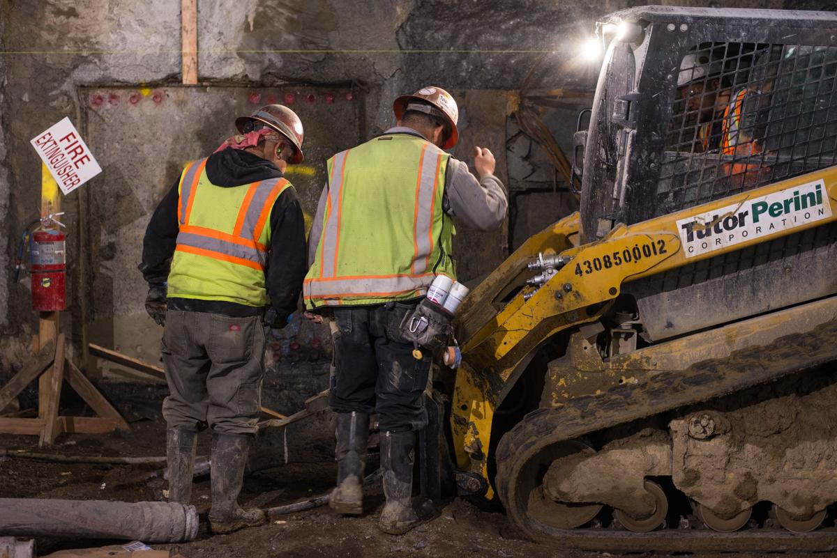 Two workers in safety vests and hard hats inspect equipment near a yellow Bobcat skid-steer loader at an underground construction site.
