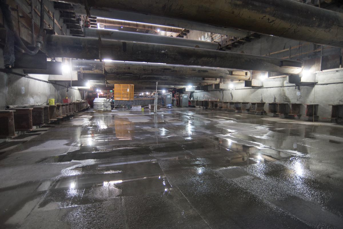 Excavated underground station box with wet concrete floor and construction lighting under overhead beams.