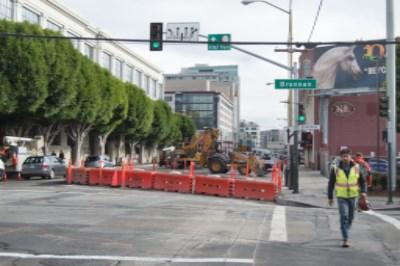 Urban street intersection with red construction barriers, traffic cones, and workers in safety vests during utility work on 4th Street.