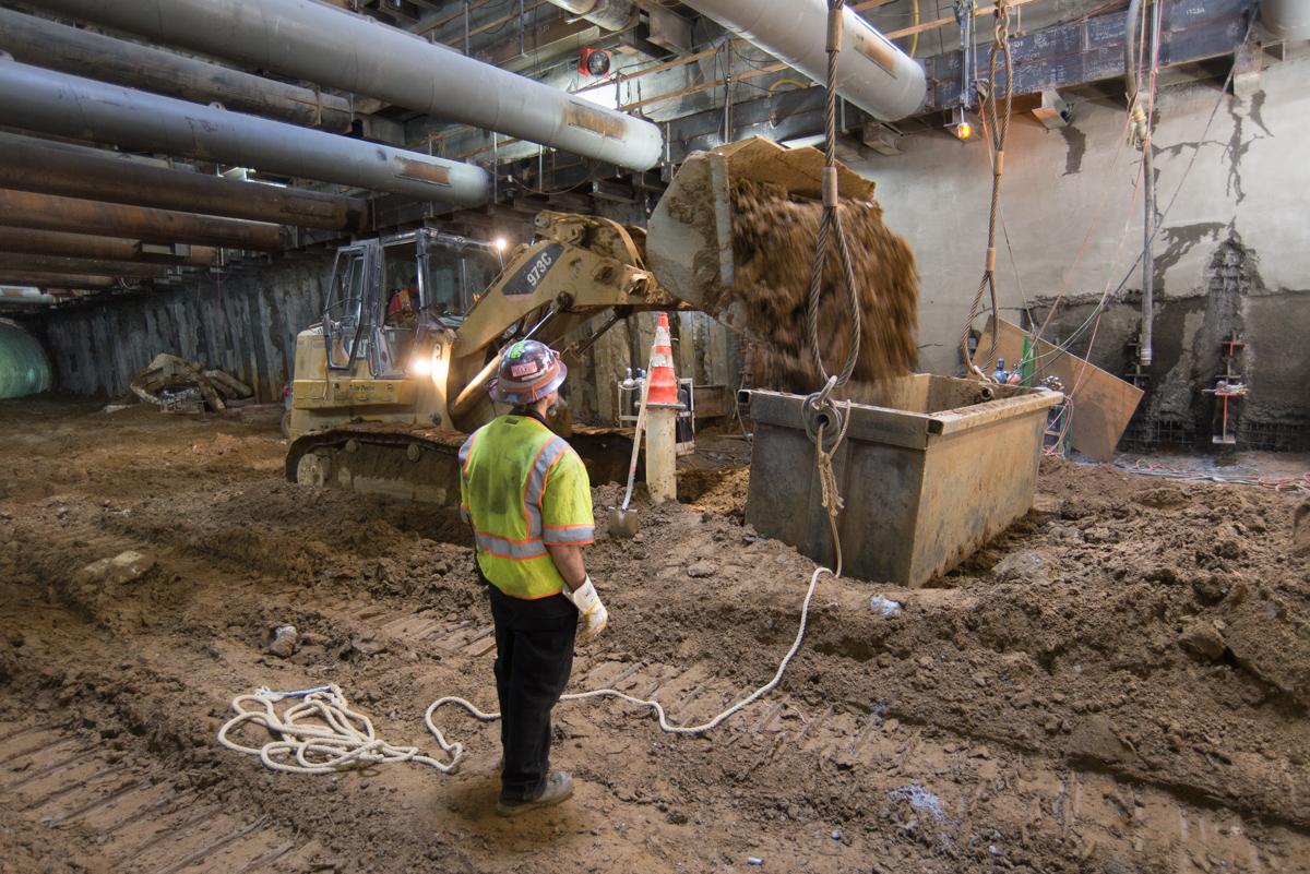 Worker in safety vest directs excavation bucket during underground station construction.