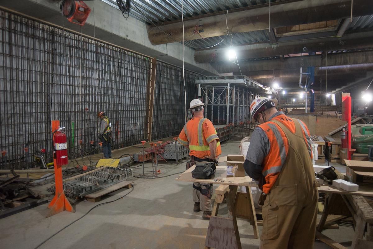 Construction workers in safety gear work inside a transit station box with concrete forms and reinforced walls.