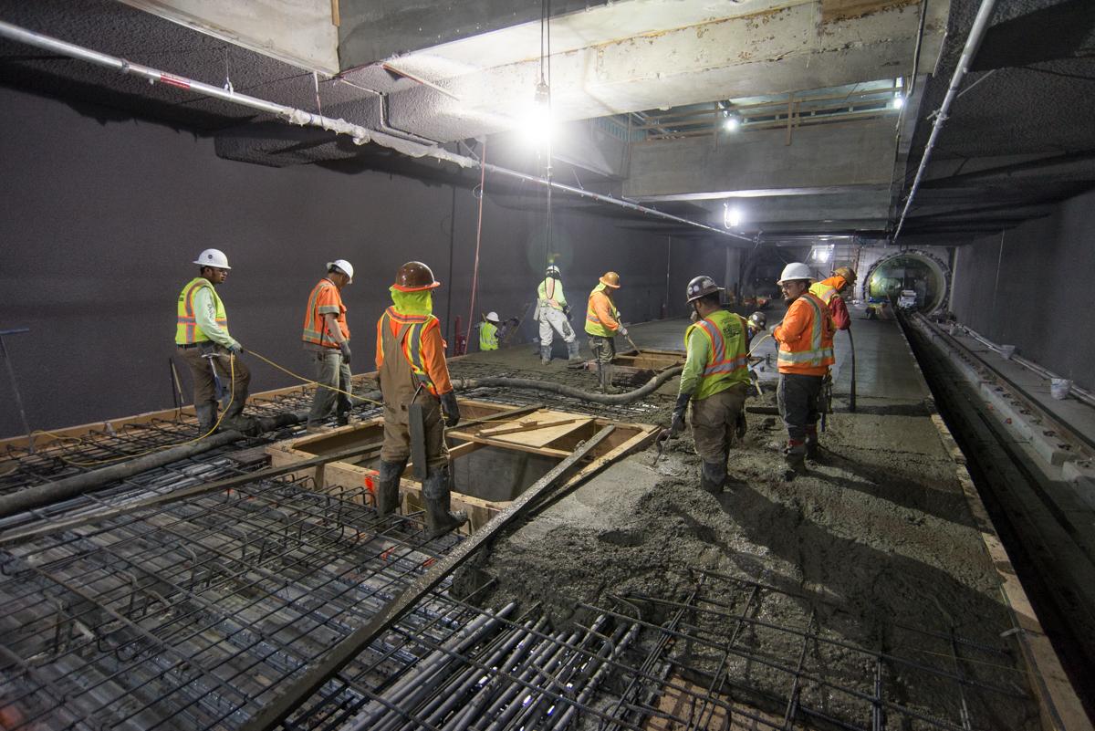 Construction workers pour concrete around elevator footings inside underground transit station box.