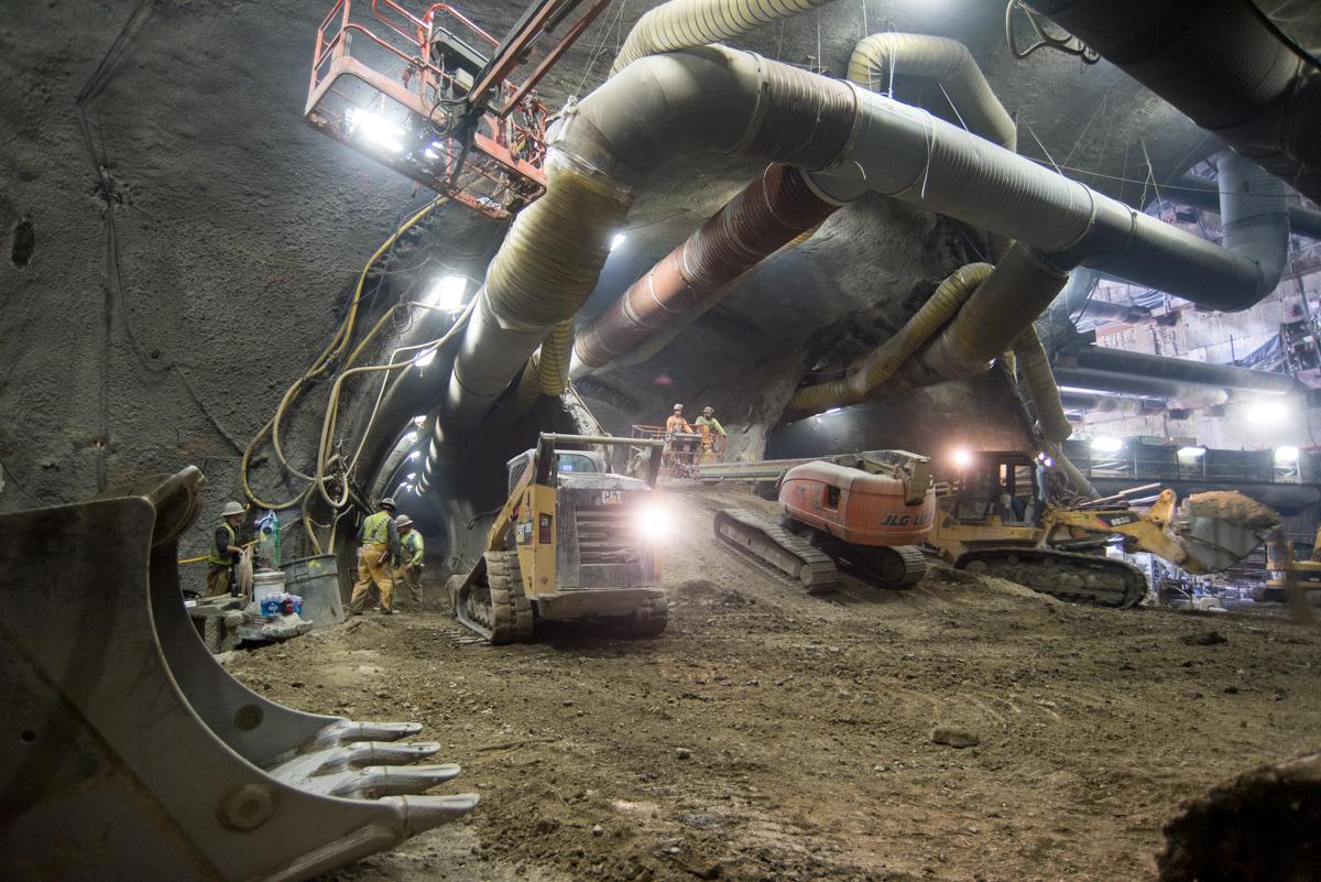 Underground tunnel construction site with large ventilation ducts, excavation equipment, and workers in safety gear.