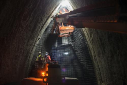 Workers install rebar in underground cavern with excavator and construction equipment visible.
