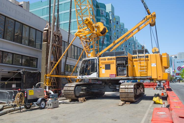 Yellow construction crane on tracked base positioned on urban street with buildings in background and red safety barriers in foreground.
