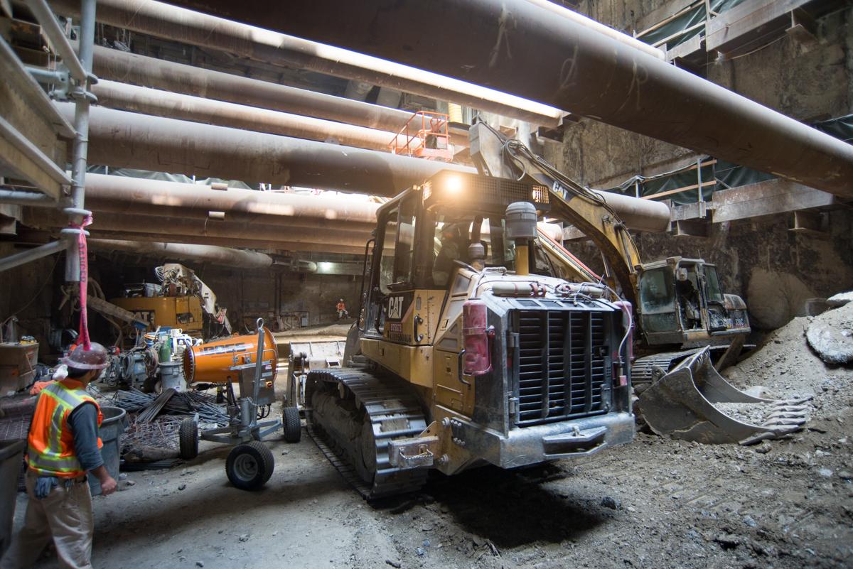 Construction equipment and workers excavate tunnel material beneath large industrial pipes at underground transit station site.