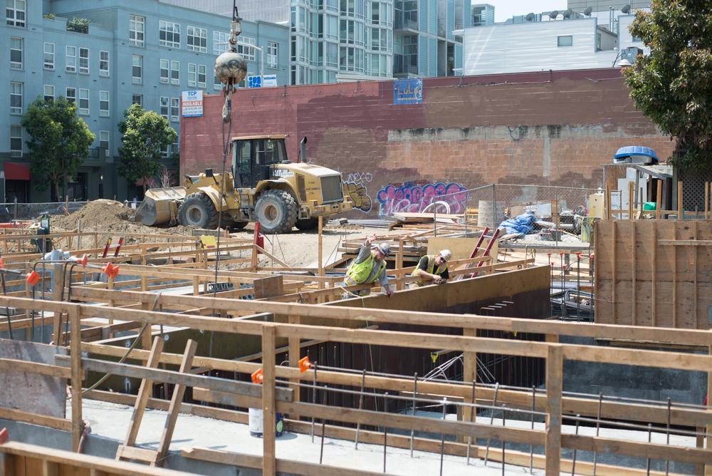 Construction site with workers, heavy equipment, and steel framework at urban transit station headhouse.