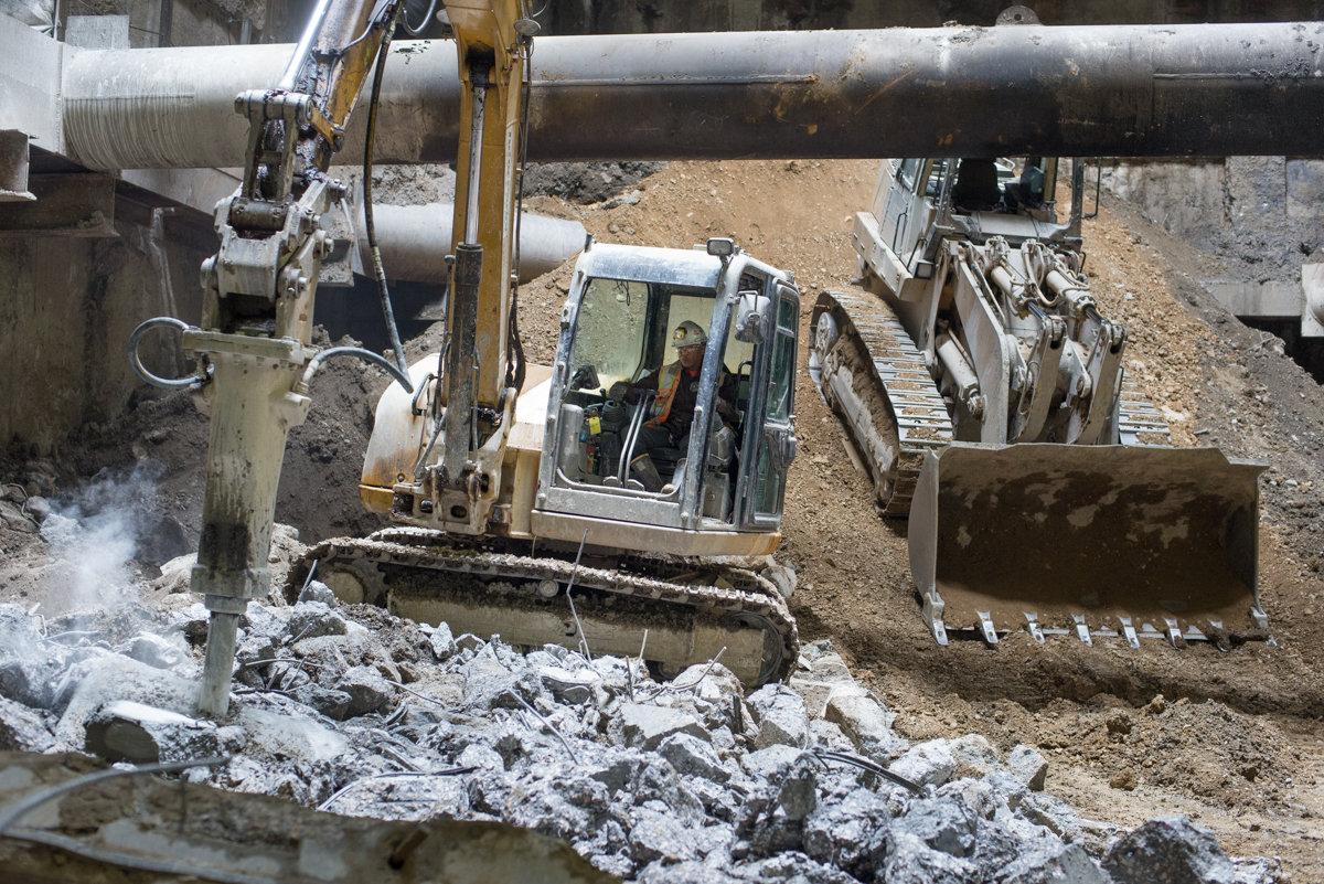 Excavator with jackhammer breaking apart concrete tunnel segments at construction site.