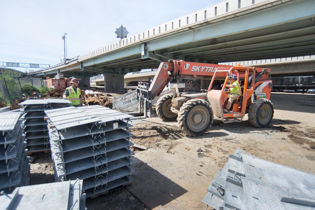 Construction equipment loads metal tunnel walkway plates at a transit project storage yard beneath an overpass.
