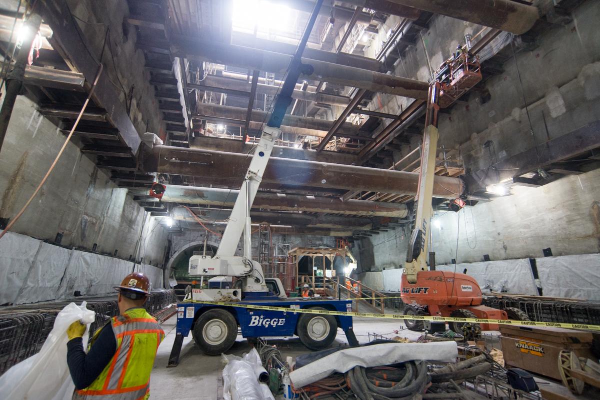 Construction workers in hard hats operate heavy equipment inside an underground tunnel, building concrete walls with steel reinforcement.