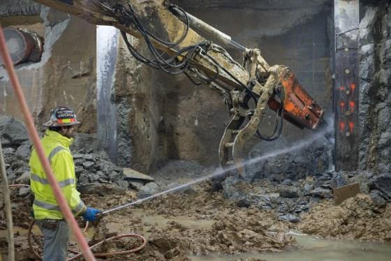 Worker in safety gear observing hydraulic demolition equipment breaking concrete in underground tunnel construction site.