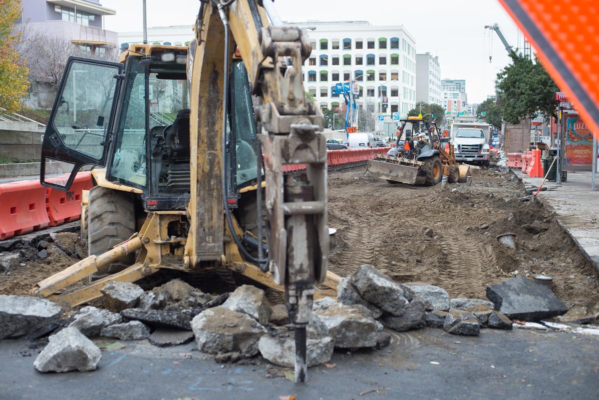 Excavator with jackhammer breaking up concrete on urban construction site near station platform.