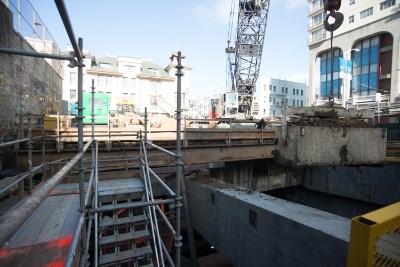 Construction site showing excavation shaft with temporary steel supports, crane, and urban buildings in background.