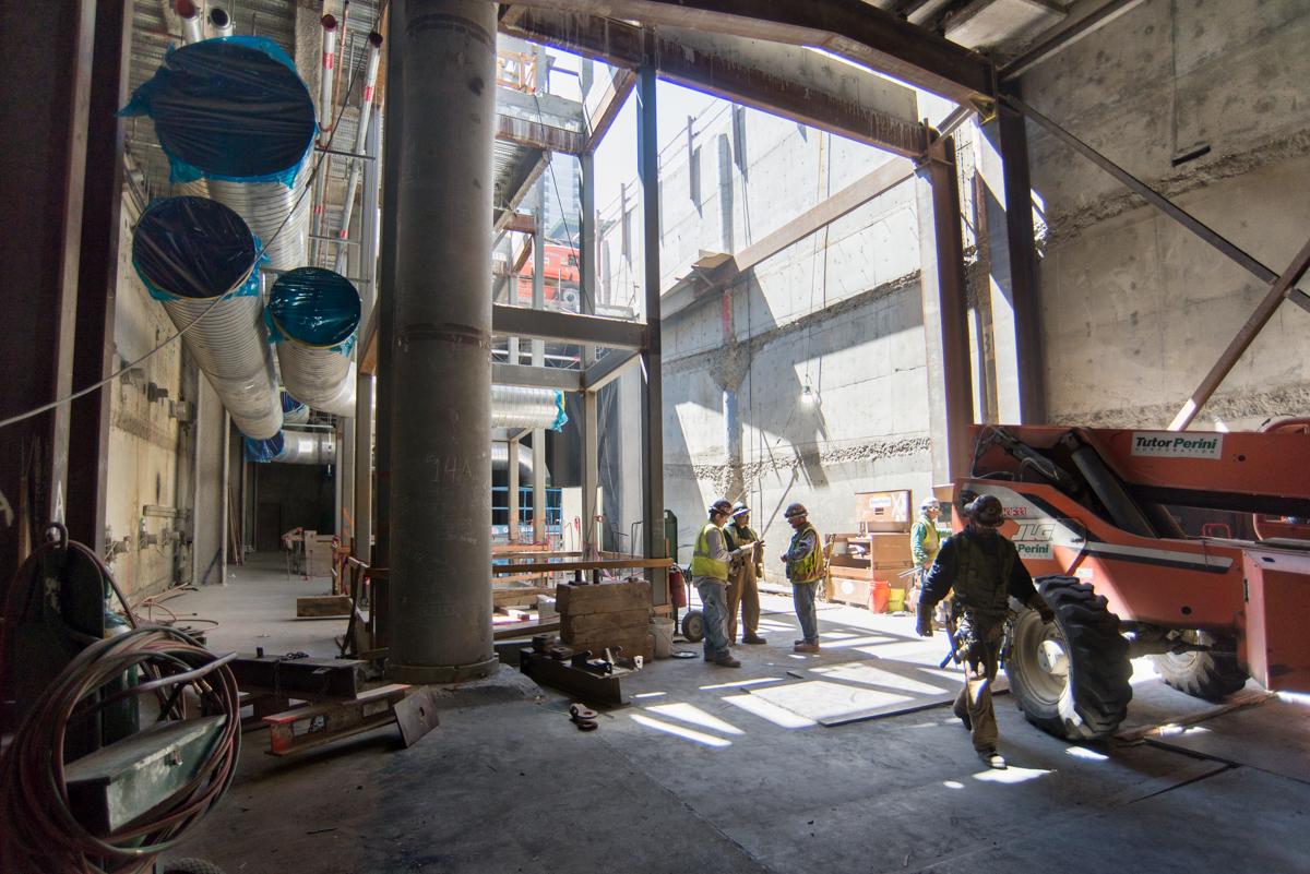 Construction workers in hard hats inside underground transit station with steel column, ventilation ducts, and equipment visible.