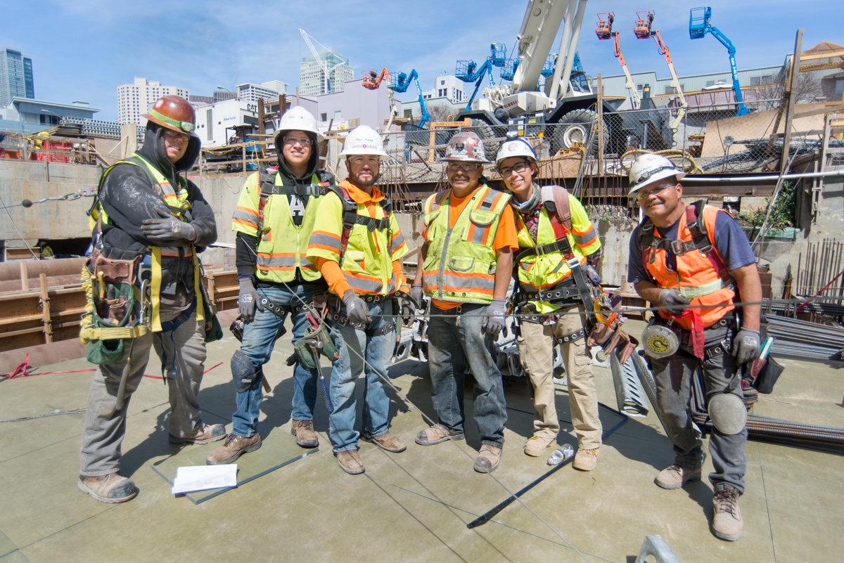 Construction crew of six workers in safety vests and hard hats posing on a transit station roof slab at an urban construction site.