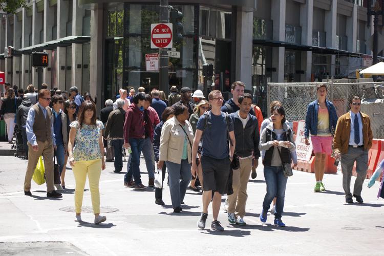 Pedestrians walking and standing on a city sidewalk near a construction site with barriers and signage.