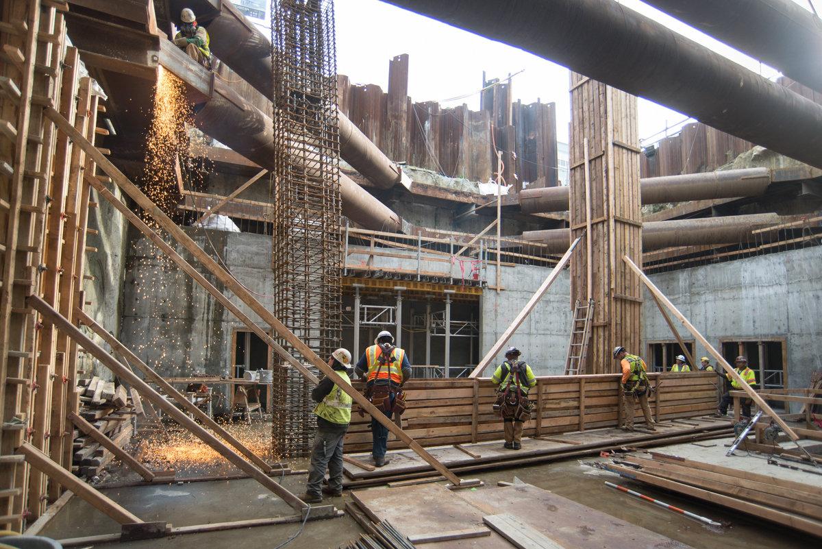 Construction workers in safety gear work on concrete formwork and steel reinforcement at an underground transit station site with city skyline visible above.