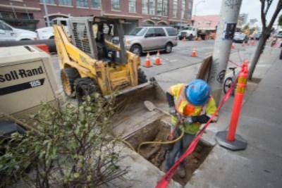 Worker with jackhammer removing concrete around utilities near open manhole and construction equipment.