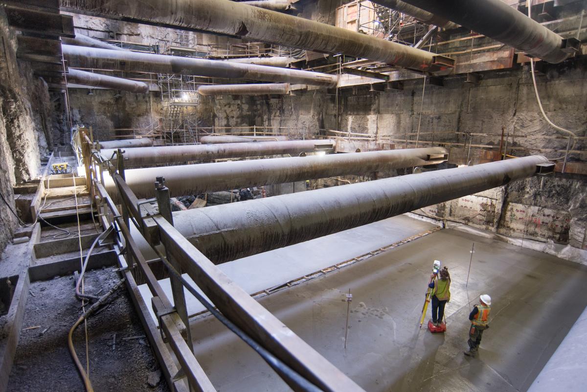Workers in safety gear survey concrete foundation at Chinatown Station construction site with large pipes overhead.