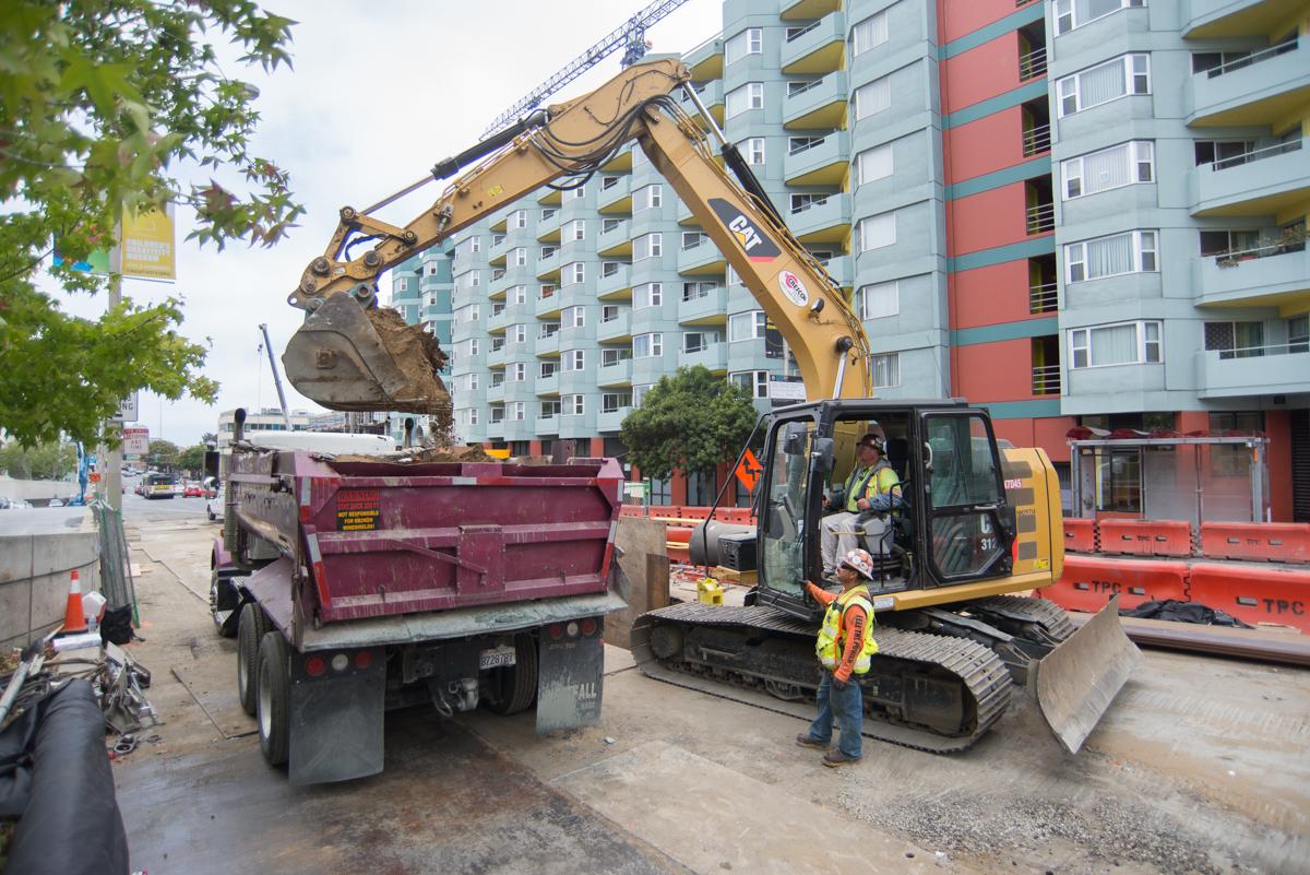 Excavator loading debris into dump truck at urban construction site near apartment buildings.