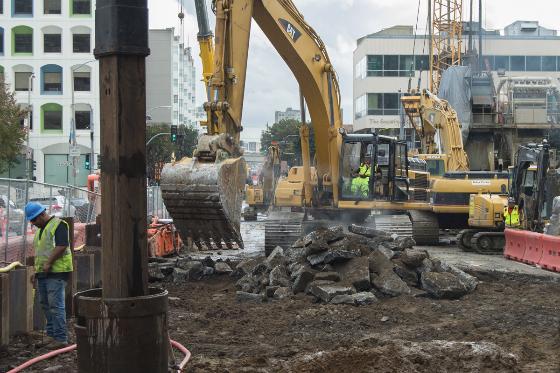 Excavator removing soil and rock at Yerba Buena/Moscone Station construction site with workers and equipment nearby.
