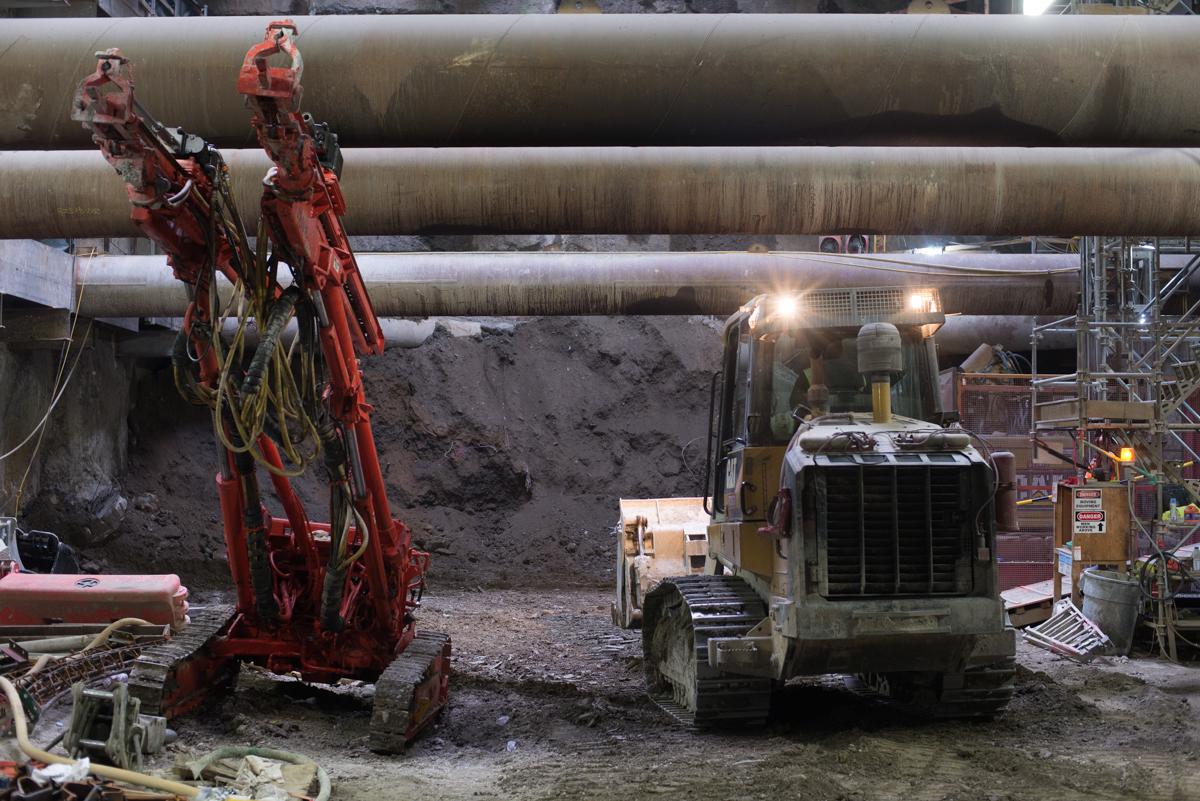 Construction site with red excavator lowering steel bucket toward bulldozer beneath large pipes.