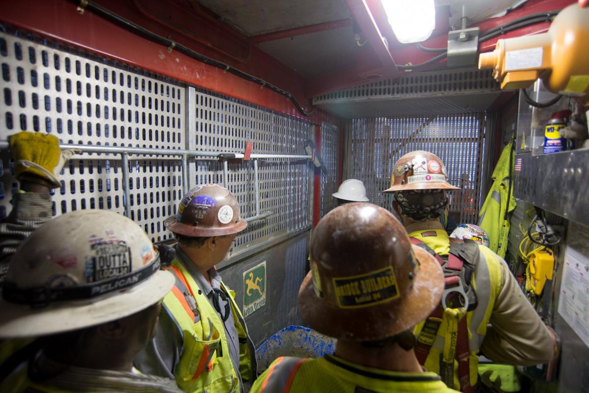 Workers in hard hats and safety vests stand in a construction elevator during station box excavation.