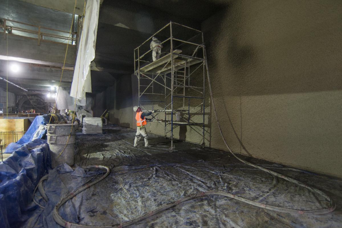 Worker in orange vest sprays sound-deadening material on station platform walls at night.