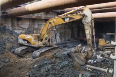 Yellow excavator operating in deep trench excavation for underground transit station construction.